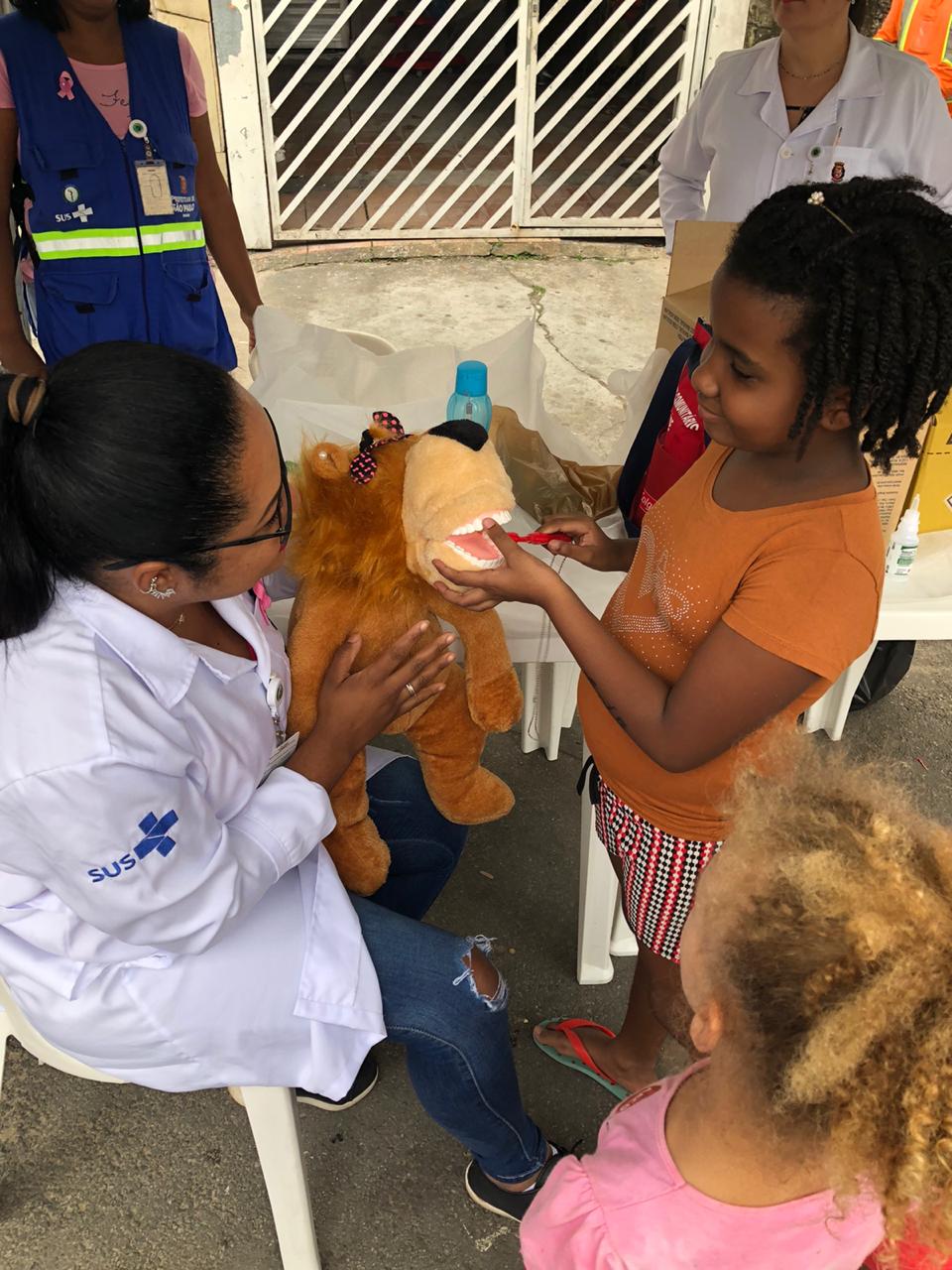 Imagem da moça do posto de saúde e 2 crianças aprendendo a escovar os dentes com um urso de pelúcia